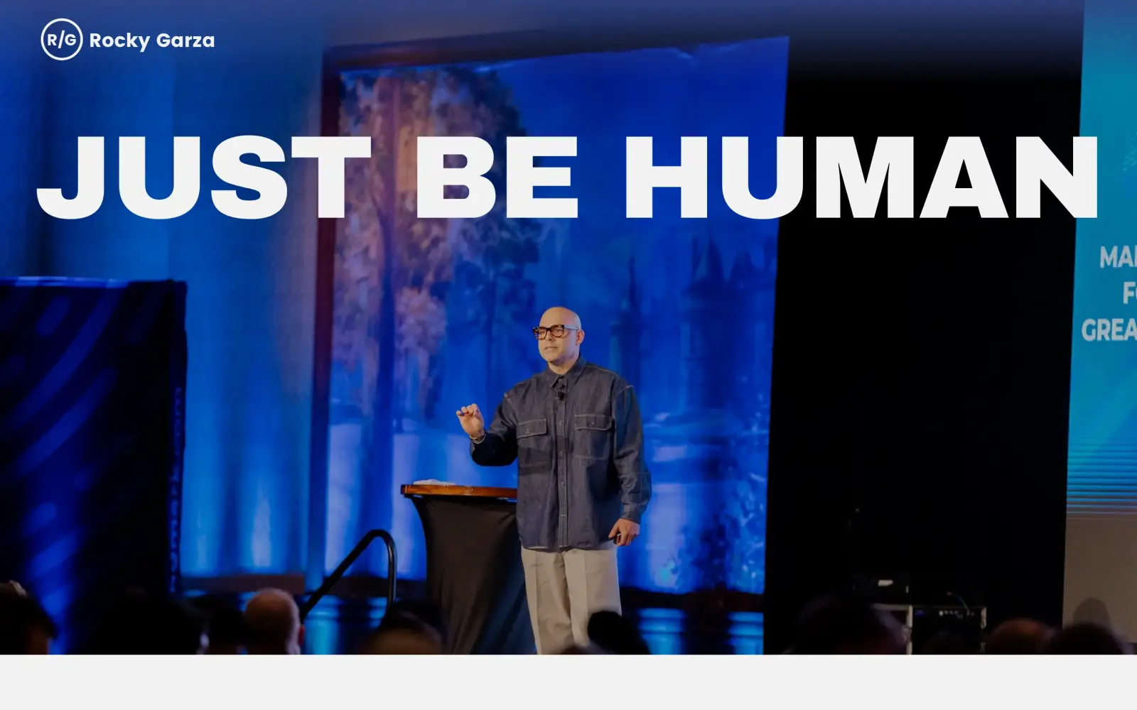 The original first screen as captured: a small R/G logo and name at top left, and the words "JUST BE HUMAN" in huge white type over a photo of Rocky speaking on a blue-lit stage. There is no menu, button or text under it.